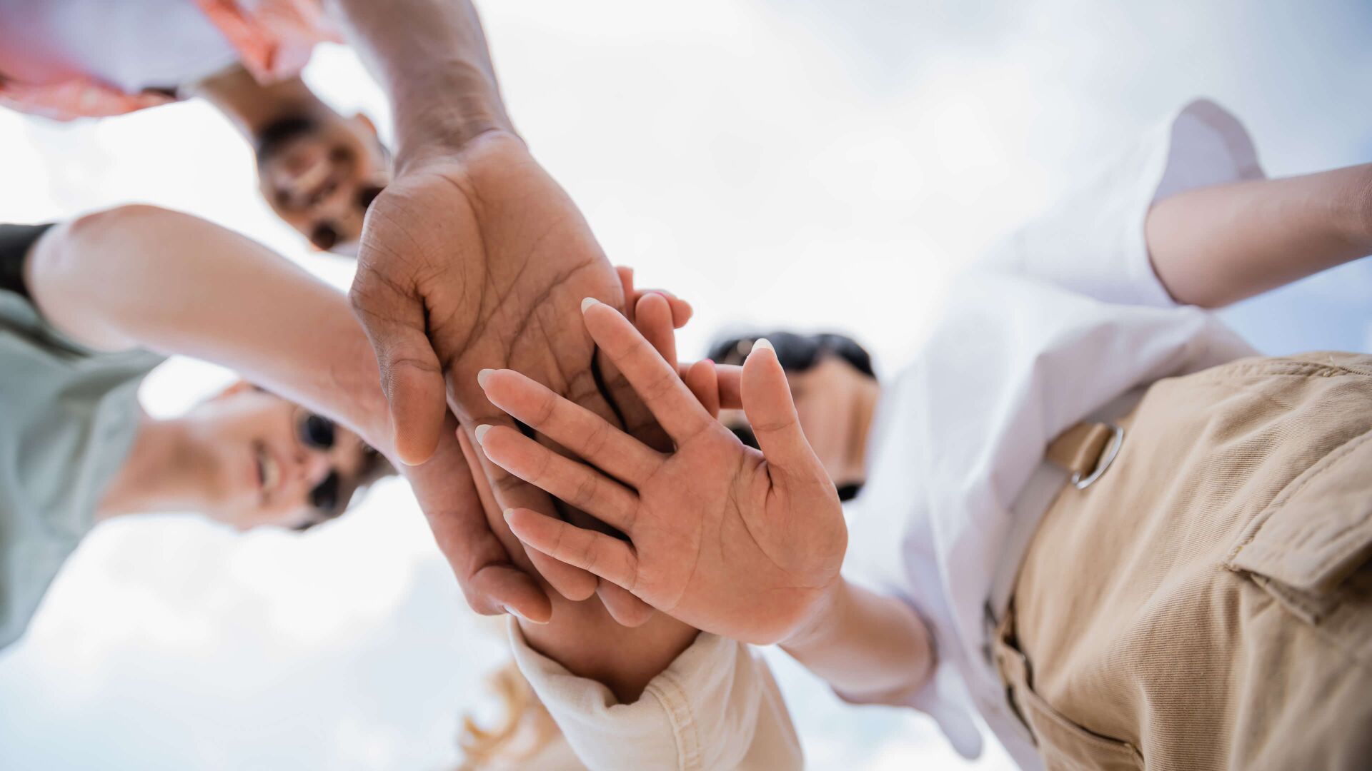 Dieses Bild zeigt vier Personen, die im Freien ihre Hände übereinanderlegen – Sinnbild für Gemeinschaft, Vertrauen und Zusammenarbeit.